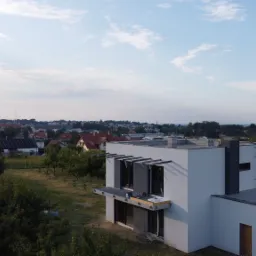 Nowoczesny, biały dom z płaskim dachem w trakcie budowy, widoczny z lotu ptaka, z elementami konstrukcyjnymi nad oknami i drabiną na dachu, na tle zabudowy miejskiej i zieleni.