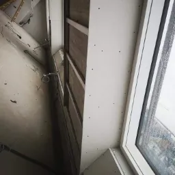 Montaż płyt gipsowo-kartonowych na ścianie z oknem, widoczne profile metalowe i ślady po wkrętach, w tle drabina i kable.