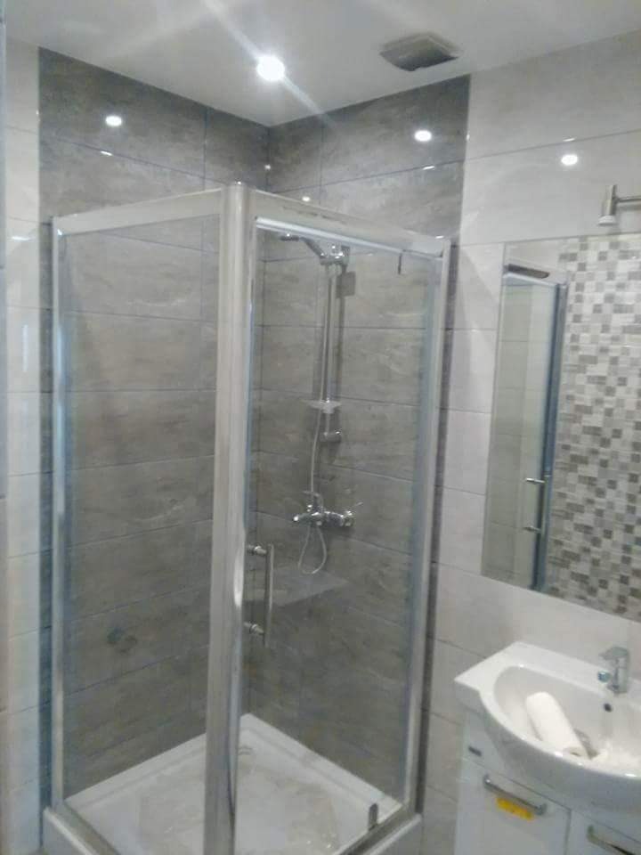 Nowoczesna kabina prysznicowa z szarymi płytkami imitującymi kamień, widoczna umywalka z szafką i lustro z mozaiką w tle.