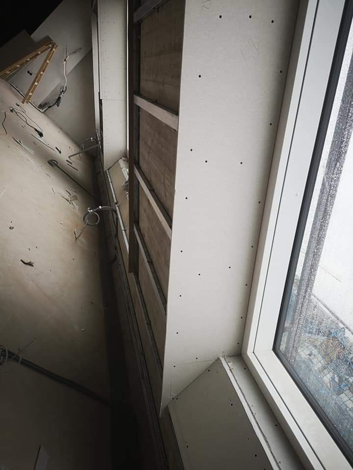 Montaż płyt gipsowo-kartonowych na ścianie z oknem, widoczne profile metalowe i ślady po wkrętach, w tle drabina i kable.