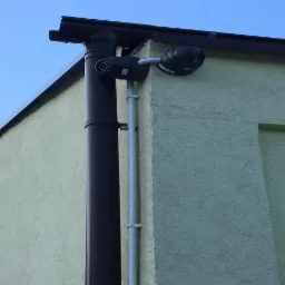 Dwie kamery monitoringu zamontowane na elewacji budynku obok rynny spustowej, widok z dołu na tle błękitnego nieba.