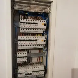 Otwarta skrzynka z bezpiecznikami, widoczne liczne wyłączniki różnicowoprądowe i przewody w różnych kolorach, zamontowana we wnęce ściany.