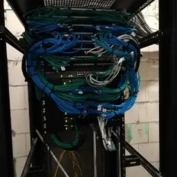 Szafa rack z routerami i chaotycznie ułożonymi kablami sieciowymi w kolorach niebieskim i zielonym, na tle surowej ściany z pustaków.