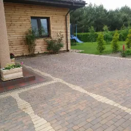 Ułożona kostka granitowa w odcieniach brązu i beżu przed drewnianym domem z kolumną, widok na trawnik z tujami i zjeżdżalnią.