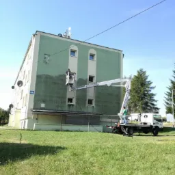 Pracownik na podnośniku koszowym czyści fragment elewacji budynku, widoczny efekt różnicy w kolorze przed i po czyszczeniu. Na pierwszym planie trawnik, w tle drzewa i błękitne niebo.