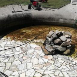 Aquadis - Czyszczenie dna fontanny z mozaiką, z użyciem myjki ciśnieniowej, na tle budynku i trawnika.