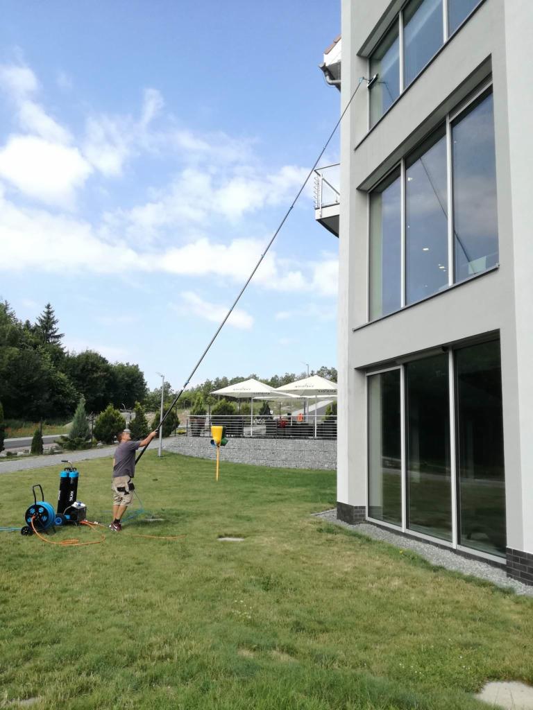 Pracownik czyści okna na wysokości za pomocą długiego lanca teleskopowego i myjki ciśnieniowej na trawniku przed budynkiem z dużymi oknami.