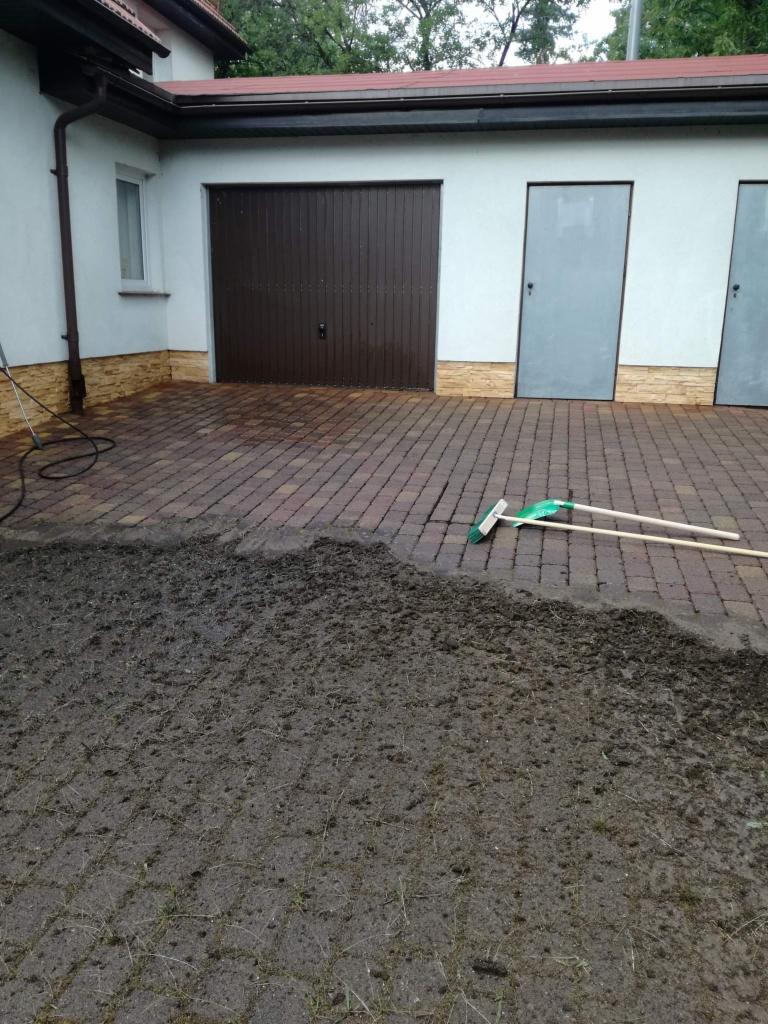 Częściowo umyta kostka brukowa przed garażem z brązową bramą, obok leży zielony miotła, widoczny brud na kostce i trawa w szczelinach.