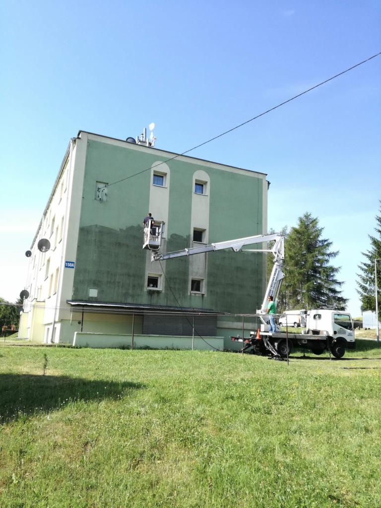 Pracownik na podnośniku koszowym czyści fragment elewacji budynku, widoczny efekt różnicy w kolorze przed i po czyszczeniu. Na pierwszym planie trawnik, w tle drzewa i błękitne niebo.