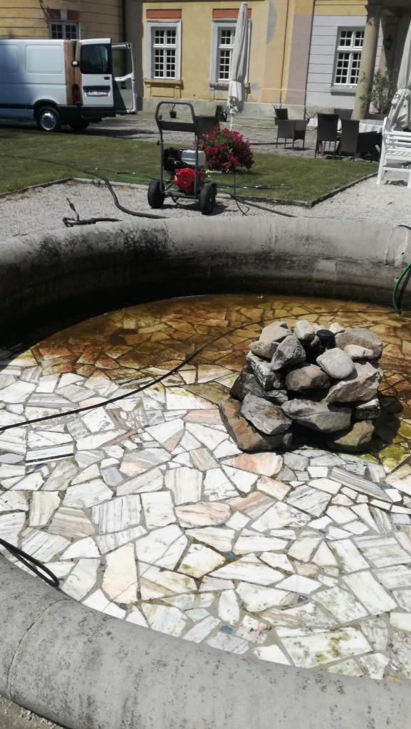 Czyszczenie dna fontanny z mozaiką, z użyciem myjki ciśnieniowej, na tle budynku i trawnika.