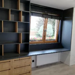 Szara zabudowa meblowa z drewnianymi akcentami obejmująca regał z otwartymi półkami, blat roboczy pod oknem i białą szafę, widok na ogród przez okno z żaluzjami.