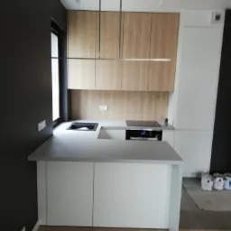 Nowoczesna kuchnia z drewnianymi szafkami górnymi, białymi dolnymi i szarym blatem, minimalistyczna lampa wisząca, ciemnoszara ściana boczna, widoczne puszki z farbą na podłodze.