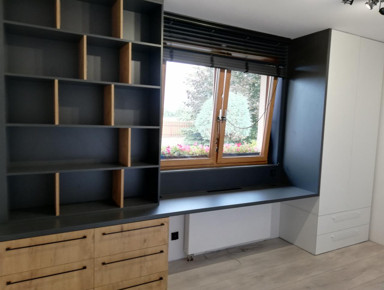 Szara zabudowa meblowa z drewnianymi akcentami obejmująca regał z otwartymi półkami, blat roboczy pod oknem i białą szafę, widok na ogród przez okno z żaluzjami.