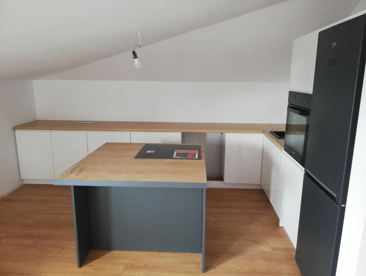 Kuchnia na poddaszu z wyspą, jasne drewniane blaty, białe szafki, czarna lodówka i piekarnik, surowy żyrandol z żarówką, widoczne niedociągnięcia wykończeniowe.