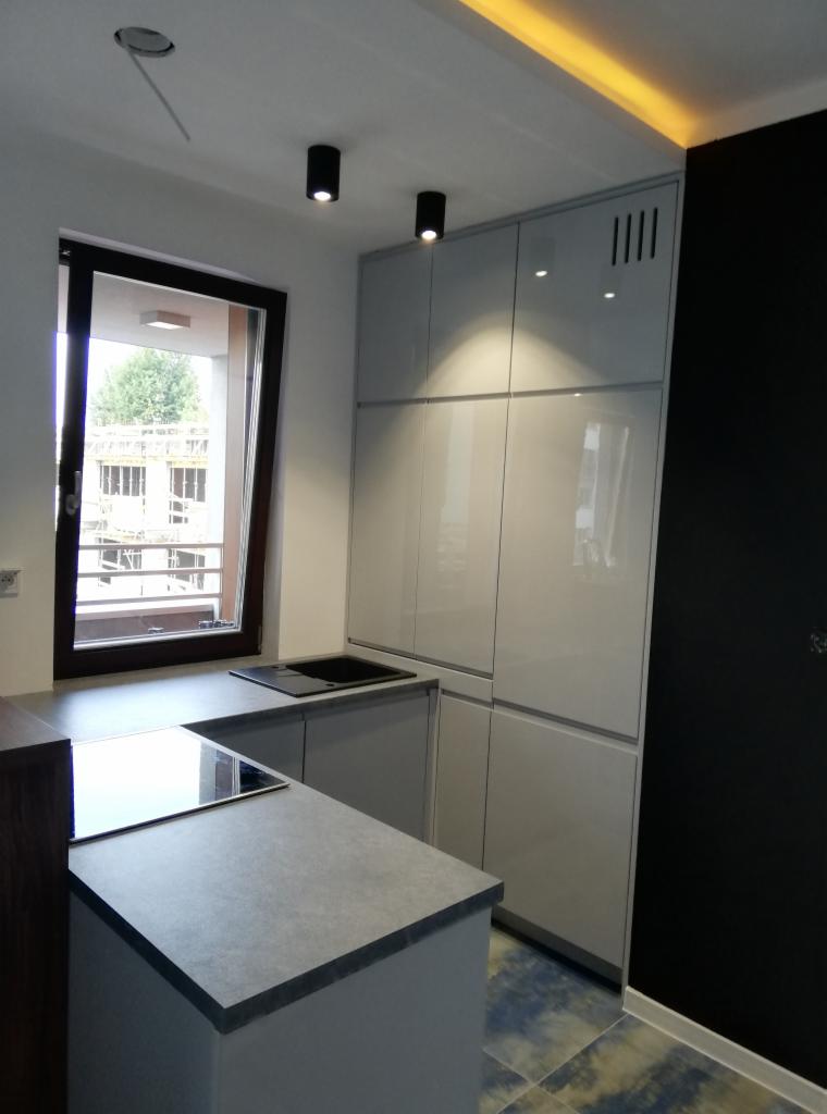 Nowoczesna, jasnoszara kuchnia z wyspą i czarnymi akcentami oświetleniowymi, widok na budynek z okna.