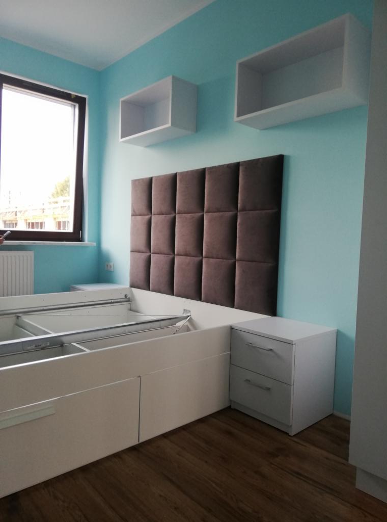 Sypialnia w trakcie montażu: białe łóżko z szufladami i stelażem, szafka nocna z dwiema szufladami, brązowy panel tapicerowany na ścianie, białe półki wiszące, błękitne ściany, drewniana podłoga.