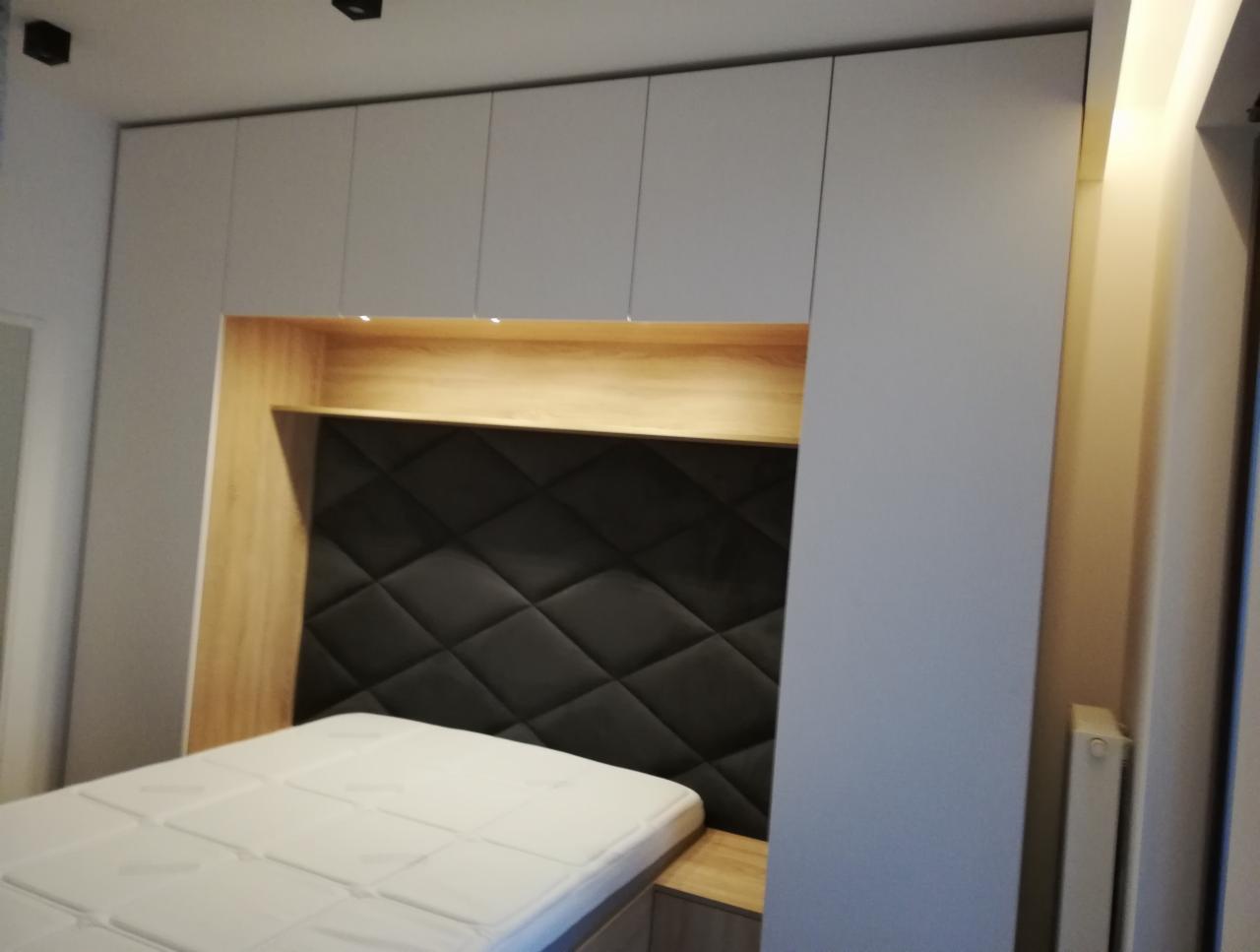 Sypialnia z zabudową: białe szafki nad łóżkiem z drewnianą wnęką i czarnym, pikowanym zagłówkiem. Widoczny fragment materaca.