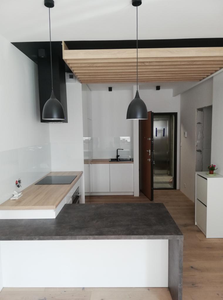 Nowoczesna kuchnia z drewnianym sufitem rastrowym, czarnymi lampami wiszącymi i blatem w kolorze ciemnego kamienia, widok na przedpokój z otwartymi drzwiami.