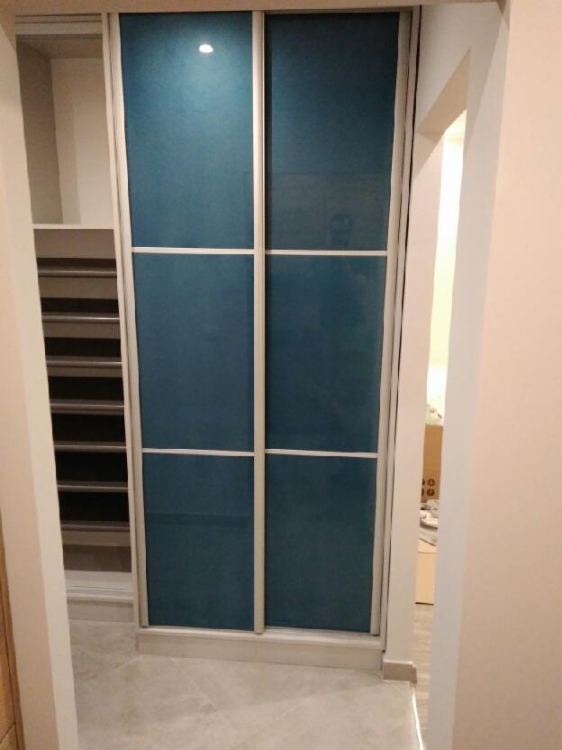 Wnęka z przesuwnymi drzwiami w kolorze ciemnego turkusu, z widocznymi półkami po lewej stronie i szarą podłogą z płytek.