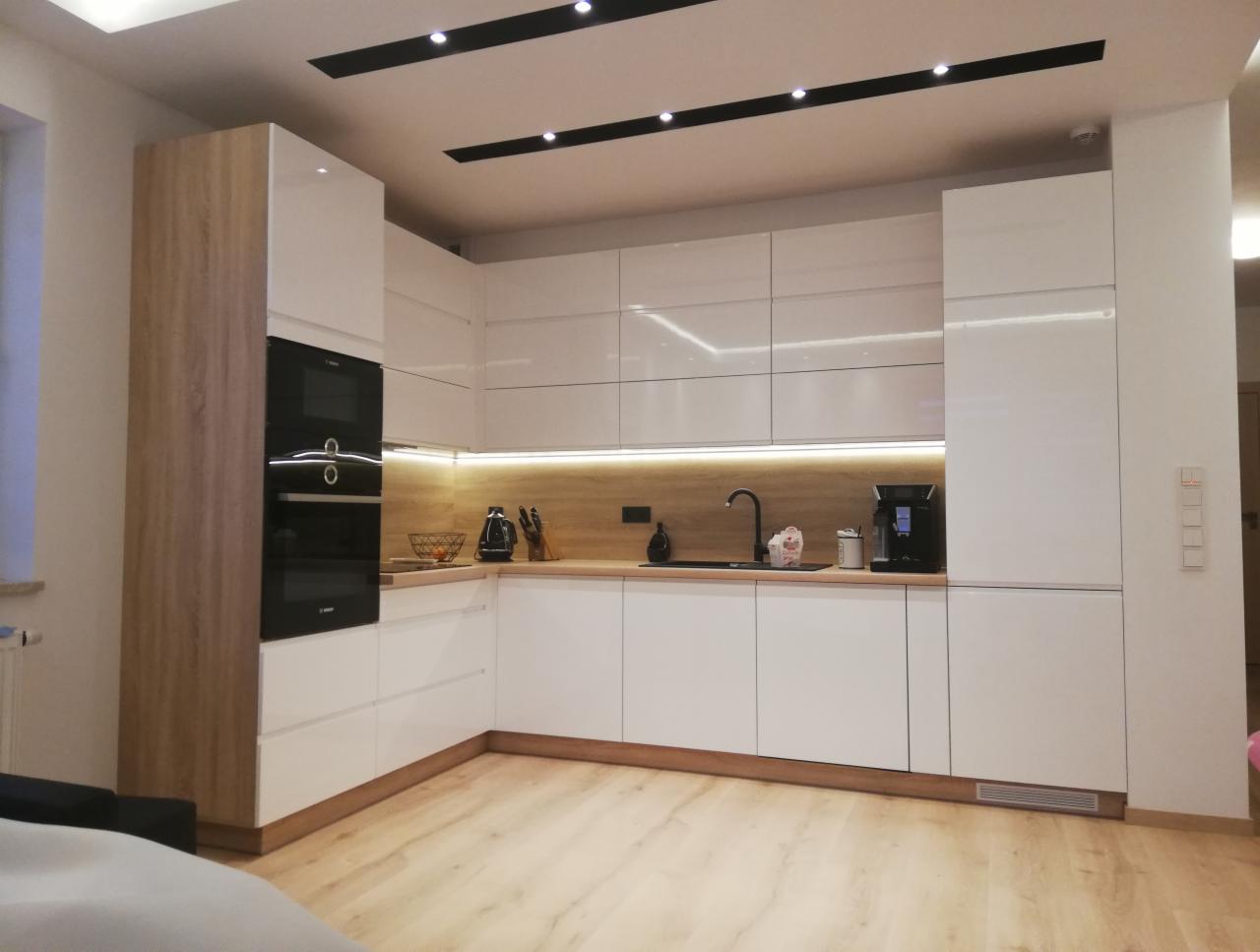 Nowoczesna kuchnia z białymi, błyszczącymi frontami szafek, drewnianym blatem i oświetleniem LED podszafkowym, z czarnymi akcentami w postaci armatury i sprzętu AGD.