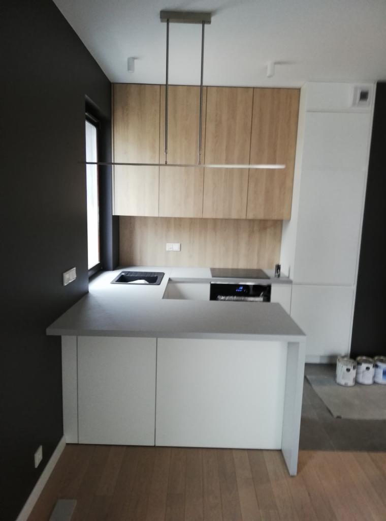 Nowoczesna kuchnia z drewnianymi szafkami górnymi, białymi dolnymi i szarym blatem, minimalistyczna lampa wisząca, ciemnoszara ściana boczna, widoczne puszki z farbą na podłodze.