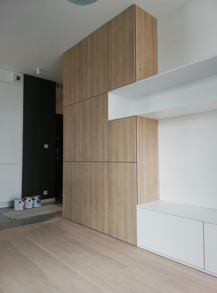 Wysoka zabudowa z drewnopodobnych paneli tworząca szafę wnękową i regał, z widocznymi puszkami farb na podłodze w trakcie prac wykończeniowych.