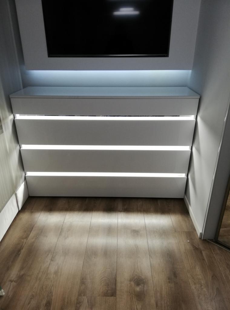 Biała komoda z czterema szufladami z podświetleniem LED wbudowana w przestrzeń pod telewizorem, na podłodze panele drewnopodobne.