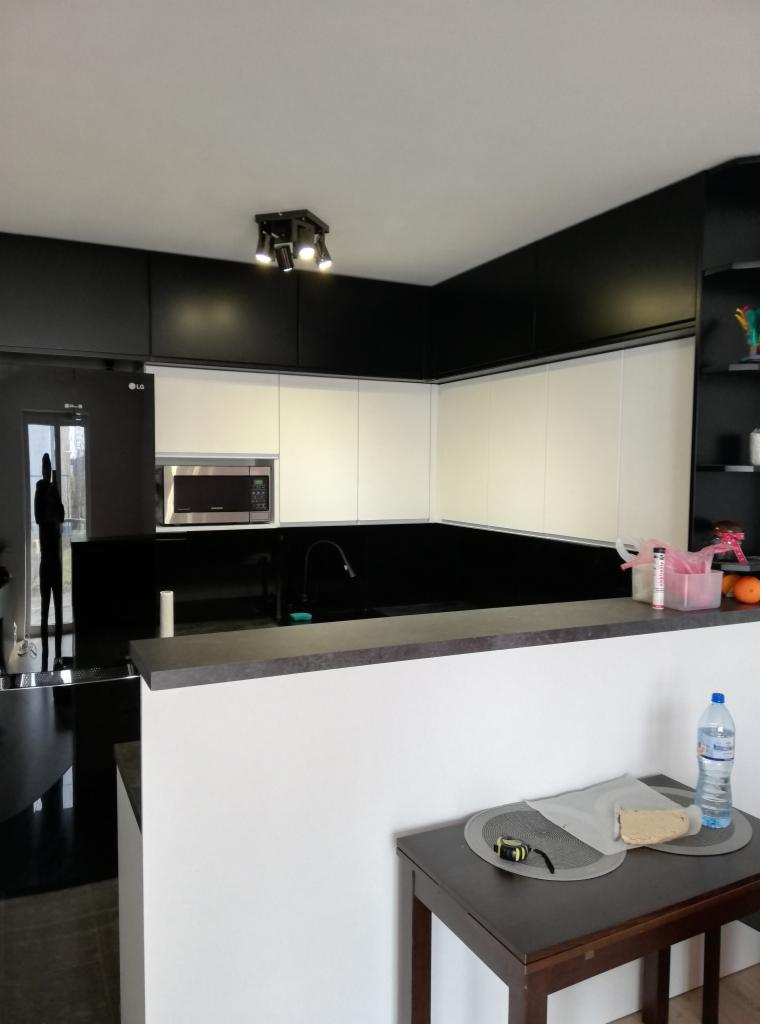 Nowoczesna kuchnia z białymi szafkami i czarnym blatem, widoczny minimalistyczny zlew, oświetlenie punktowe sufitowe, na stole w tle butelka wody i miarka.