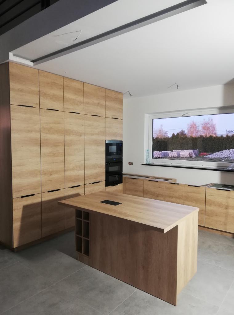 Jasna kuchnia z drewnianymi frontami, wysoka zabudowa z piekarnikiem, wyspa z półkami na wino, widok na okno z materiałami budowlanymi na zewnątrz.