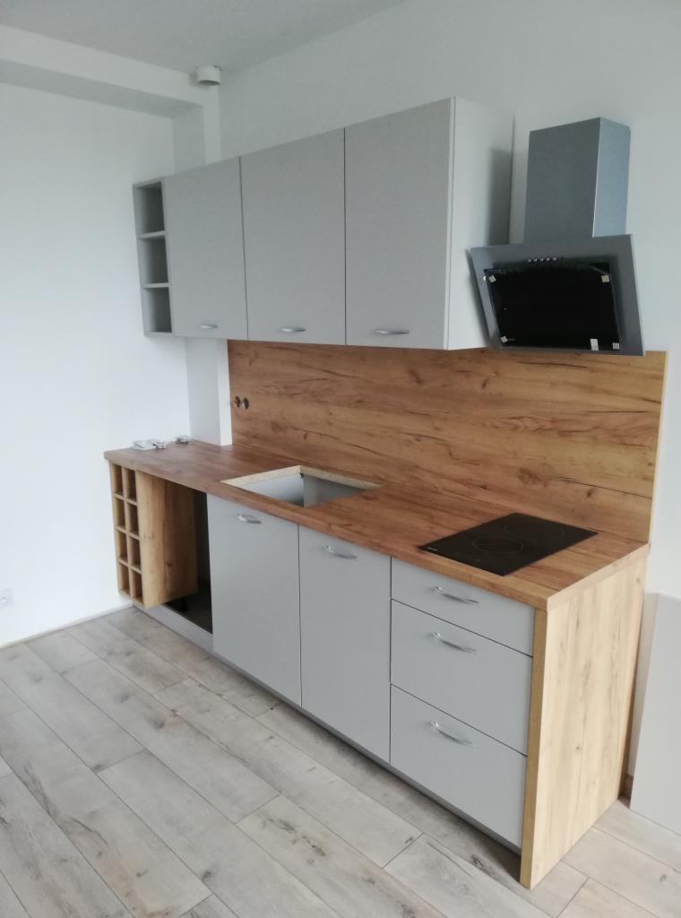 Nowoczesna kuchnia w odcieniach szarości z drewnianym blatem i backsplashiem, widoczny zlew bez baterii oraz płyta indukcyjna, uzupełniona o szafki górne i okap.