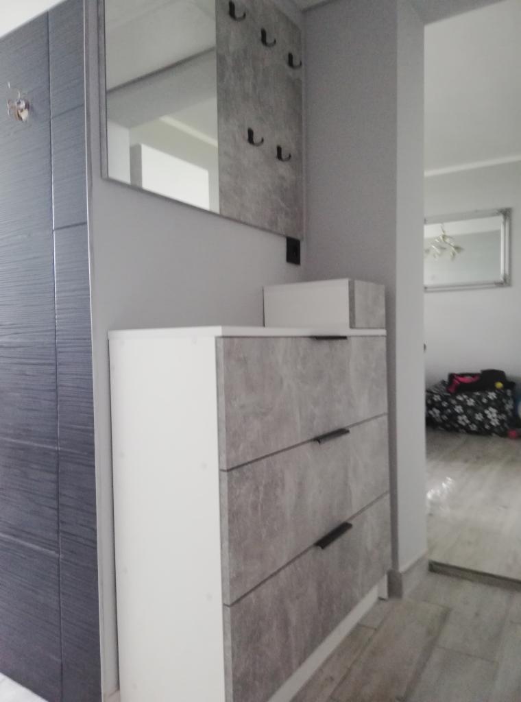 Szara szafka z trzema szufladami o marmurowym wzorze, uzupełniona lustrem z wieszakami w przedpokoju, widok z boku.