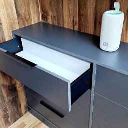 ABT Service Meble - Szara komoda z wysuniętą szufladą z białym wnętrzem, na blacie głośnik Bluetooth, tło z drewnianych paneli.