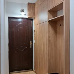 Przedpokój z zabudowaną szafą w odcieniach drewna, z siedziskiem i półkami, widoczne drzwi wejściowe i logo firmy na górze kadru.