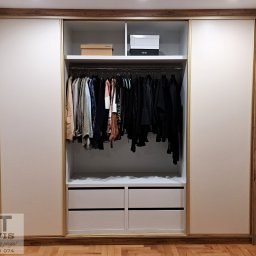 ABT Service Meble - Jasna szafa wnękowa z przesuwnymi drzwiami, drewnianą ramą i złotymi akcentami. Wnętrze z półkami, drążkiem na ubrania i szufladami. Widoczny parkiet na podłodze.