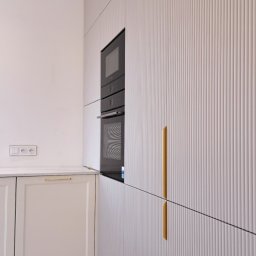 ABT Service Meble - Nowoczesna kuchnia z frezowanymi frontami w odcieniach bieli, złote uchwyty, zabudowa piekarnika i płyty grzewczej. Minimalistyczny design, subtelne oświetlenie.