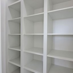 ABT Service Meble - Biała, pusta szafa wnękowa z półkami i szufladami, oświetlenie LED u góry, widok z dołu, logo firmy w prawym górnym rogu. Minimalistyczny design.