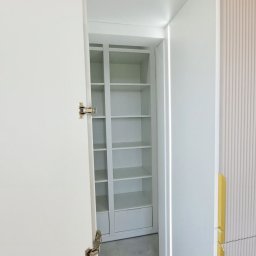 ABT Service Meble - Otwarta, biała szafa wnękowa z półkami, widoczna z bliska. Uwagę zwracają metalowe zawiasy i minimalistyczny design. Tło stanowi ściana z delikatnym, pionowym wzorem.