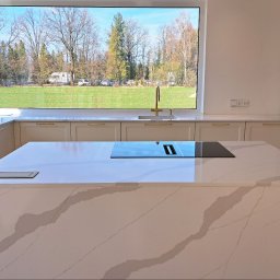 ABT Service Meble - Elegancka kuchnia z wyspą i lakierowanymi frontami w odcieniach kaszmiru. Blaty ze spieku kwarcowego, złote uchwyty. Duże okno z widokiem na zieleń.