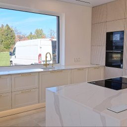 ABT Service Meble - Elegancka kuchnia z wyspą i złotymi akcentami. Widok na zewnątrz przez duże okno, gdzie stoi biały bus. Fronty szafek kremowe, ryflowane. Nowoczesny design.