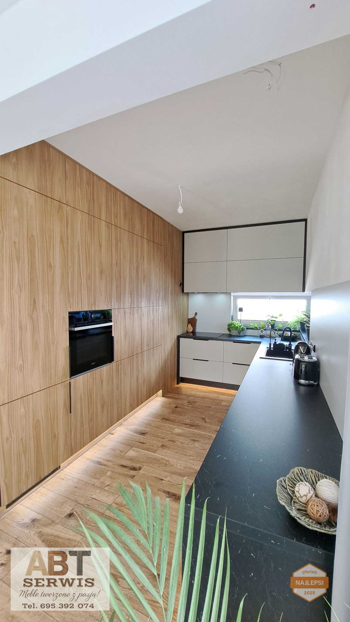 Nowoczesna kuchnia na wymiar z drewnianymi frontami i czarnym blatem, oświetlenie LED pod szafkami, roślina w tle, wąskie ujęcie perspektywiczne.