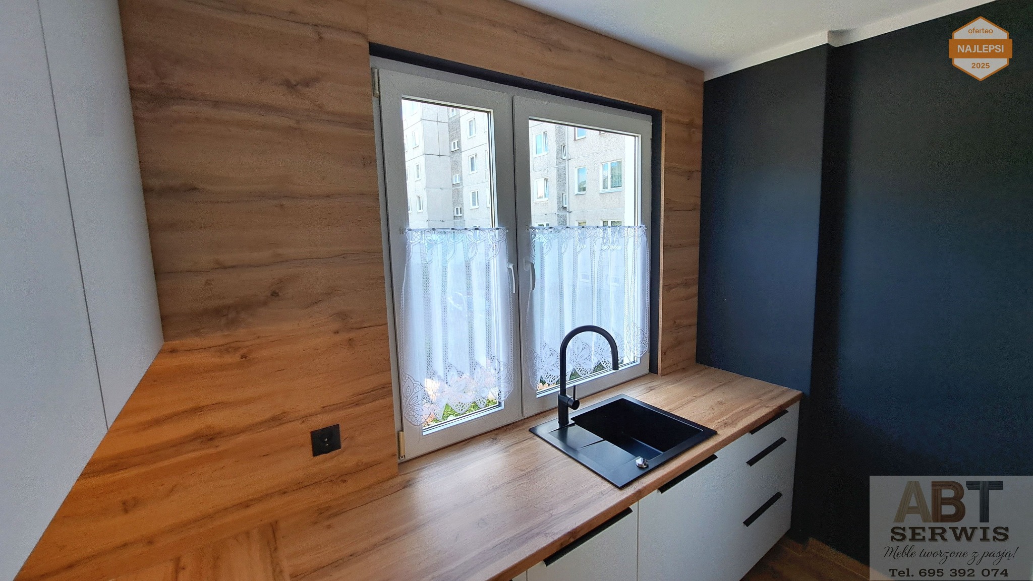 Nowoczesna kuchnia z drewnianym blatem i czarnym zlewem pod oknem z firanką. Ściana w kolorze grafitowym. Widoczne logo firmy ABT Serwis.