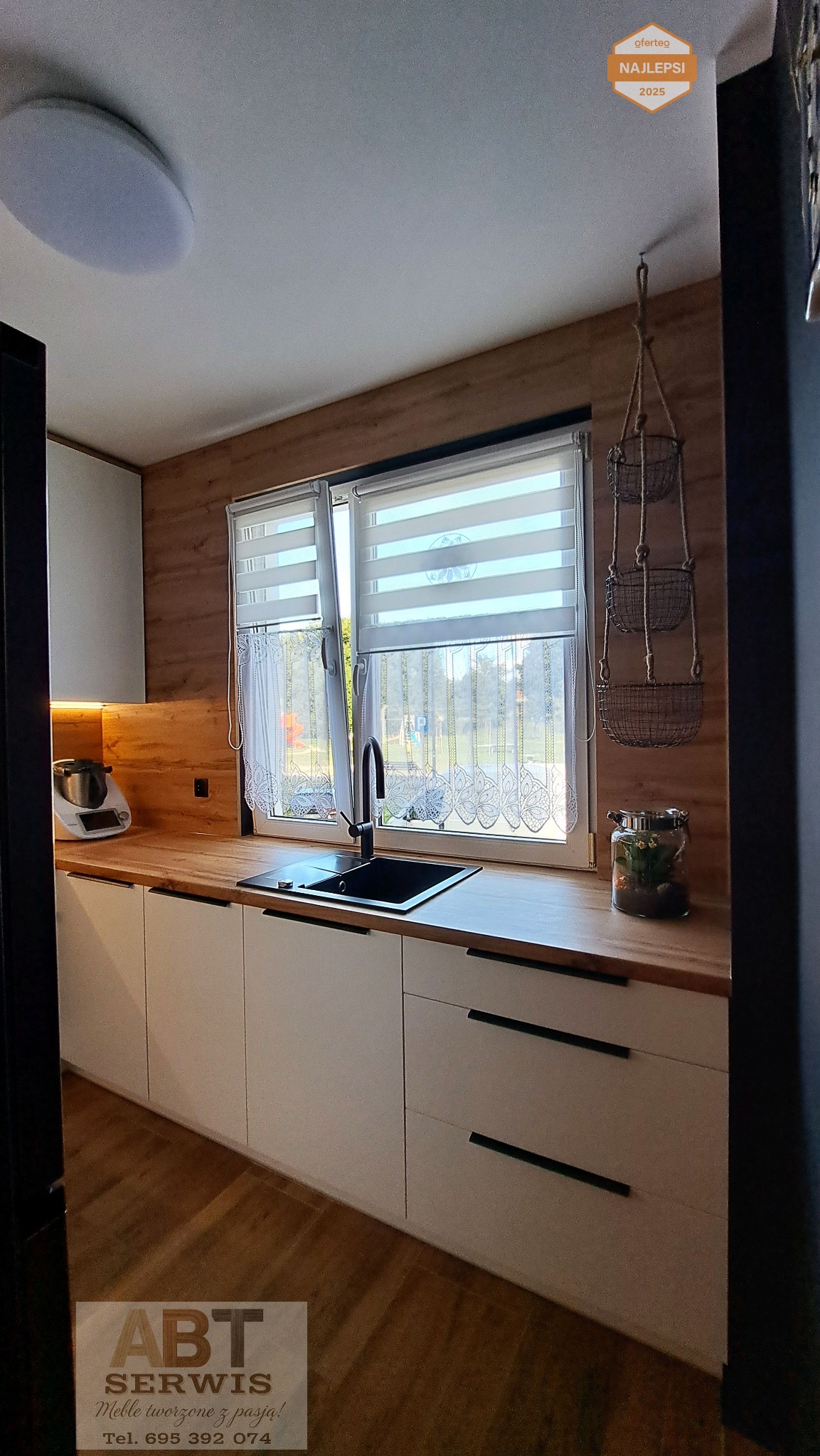 Jasna kuchnia z drewnianym blatem i czarnymi uchwytami, okno z firanką i roletami dzień-noc. Uwagę przyciąga wisząca dekoracja z koszyków.