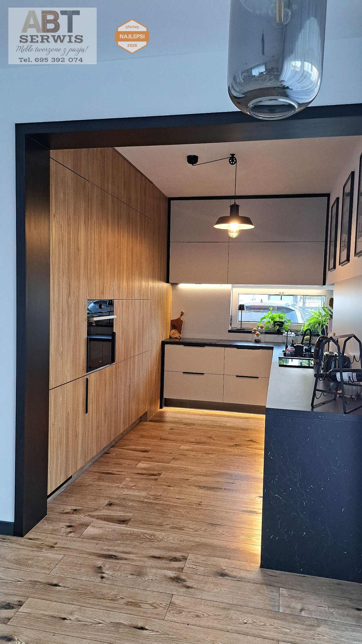 Nowoczesna kuchnia z drewnianymi frontami szafek, czarnymi akcentami i oświetleniem LED pod szafkami. Widoczny piekarnik w zabudowie i drewniana podłoga.