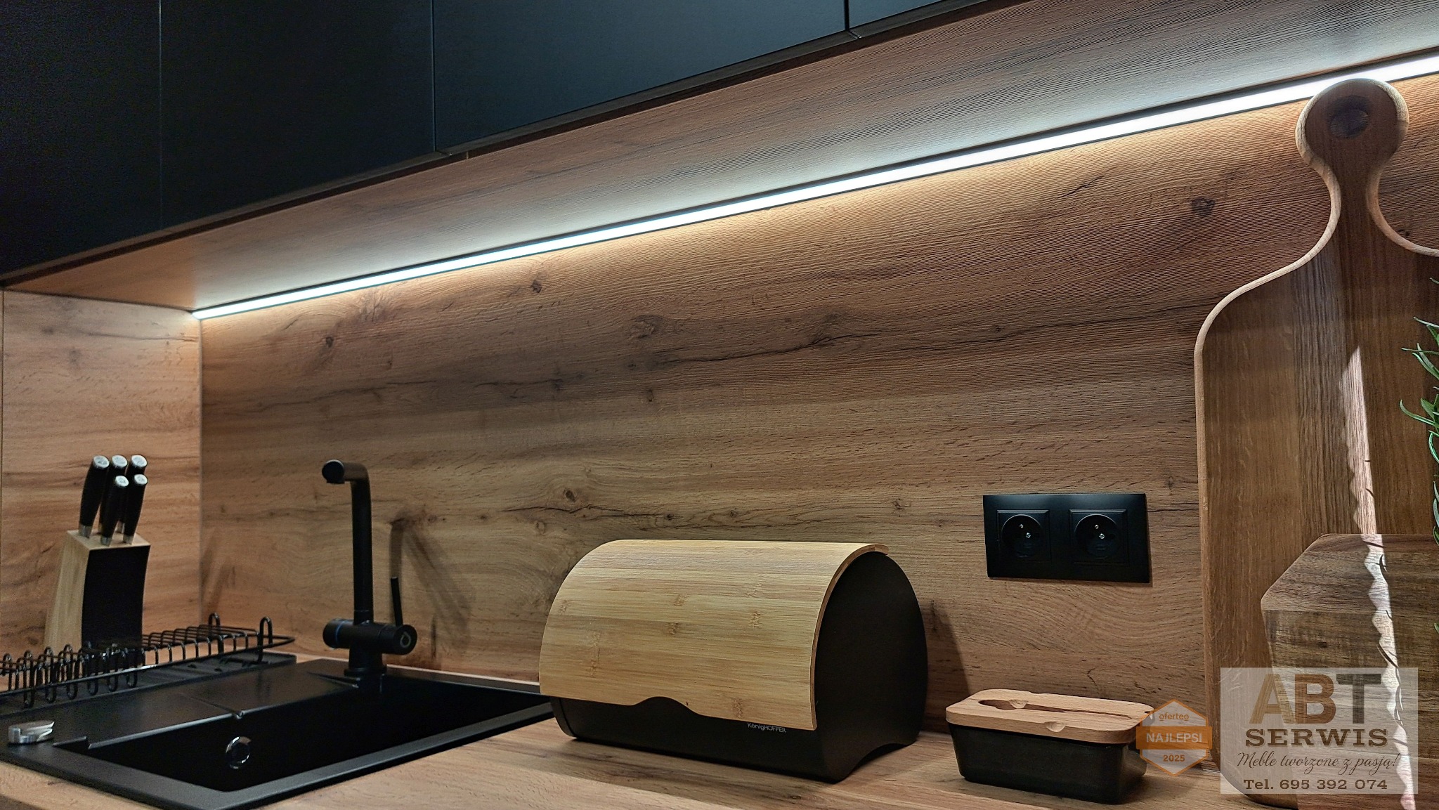 Nowoczesna kuchnia z drewnianym blatem i podświetleniem LED. Czarny zlew, ociekacz i akcesoria. Na blacie chlebak i deska do krojenia.