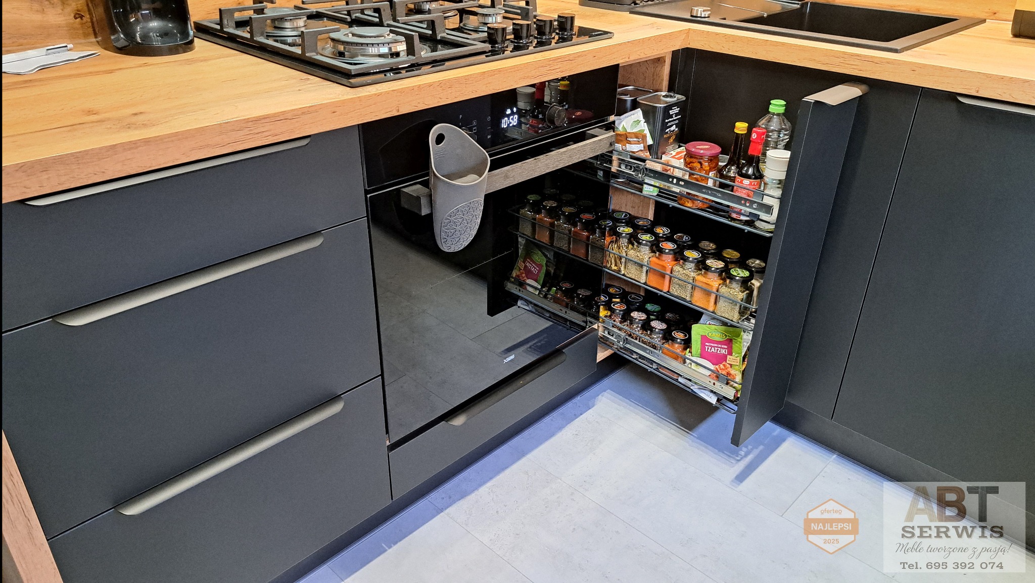 Ciemna kuchnia z drewnianym blatem. Wysuwana szafka cargo na przyprawy obok piekarnika. Organizer na drobiazgi zawieszony na piekarniku. Widoczny fragment płyty grzewczej.