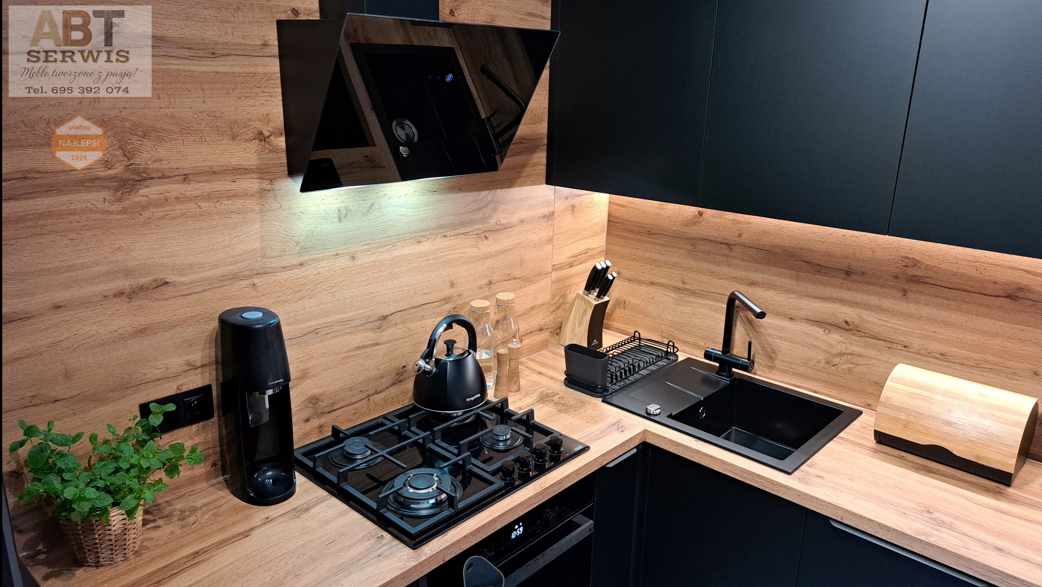 Nowoczesna kuchnia z czarnymi szafkami i drewnianym blatem. Na blacie czajnik, płyta gazowa, zlew i akcesoria kuchenne. Nad blatem czarny okap.