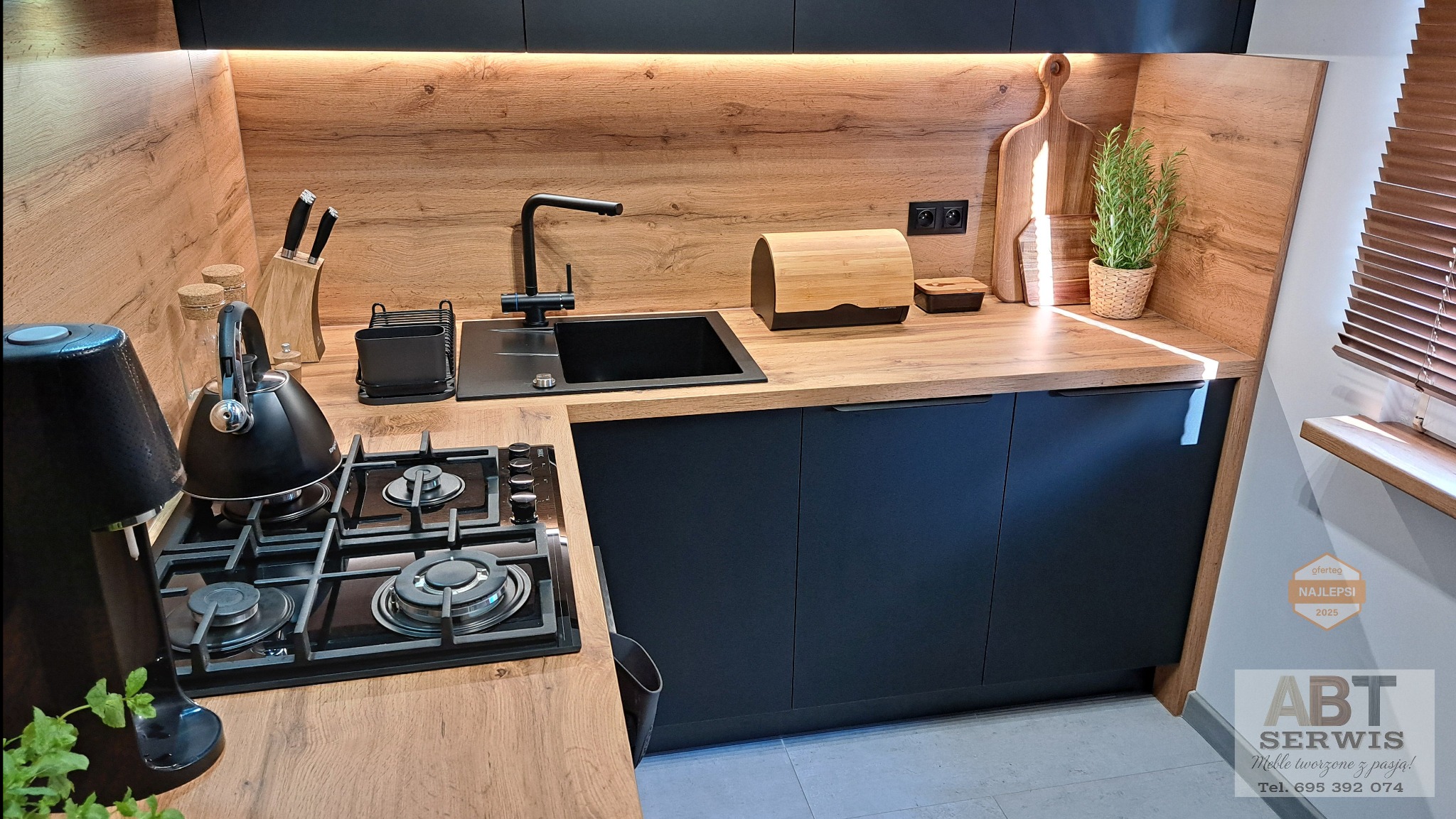 Nowoczesna kuchnia z czarnymi szafkami i drewnianym blatem. Widoczny zlew, płyta grzewcza i akcesoria. Ciepłe oświetlenie podkreśla fakturę drewna.