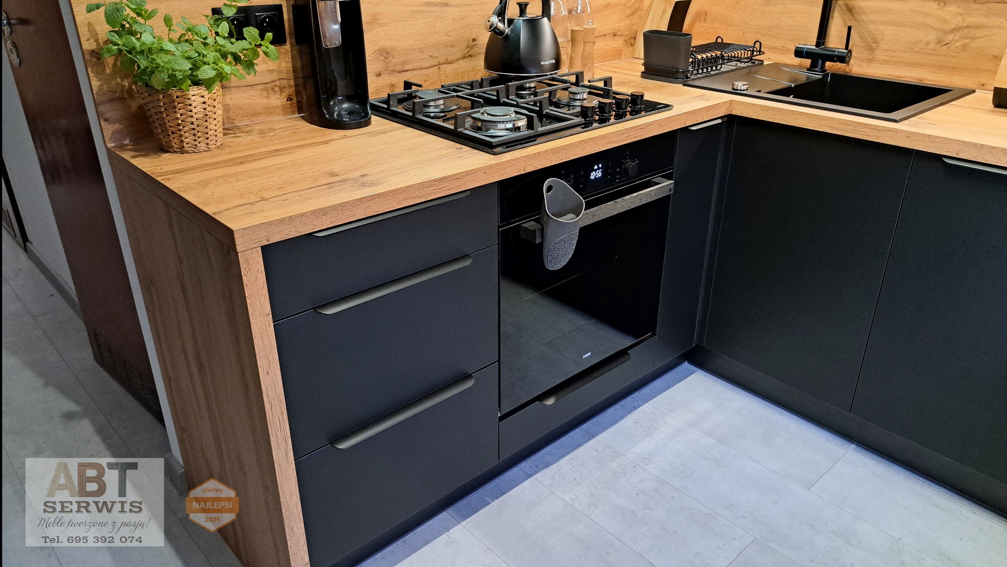 Nowoczesna kuchnia na wymiar z czarnymi frontami i drewnianym blatem. Widoczna płyta grzewcza, piekarnik i zlewozmywak. Minimalistyczny design z akcentami roślinnymi.