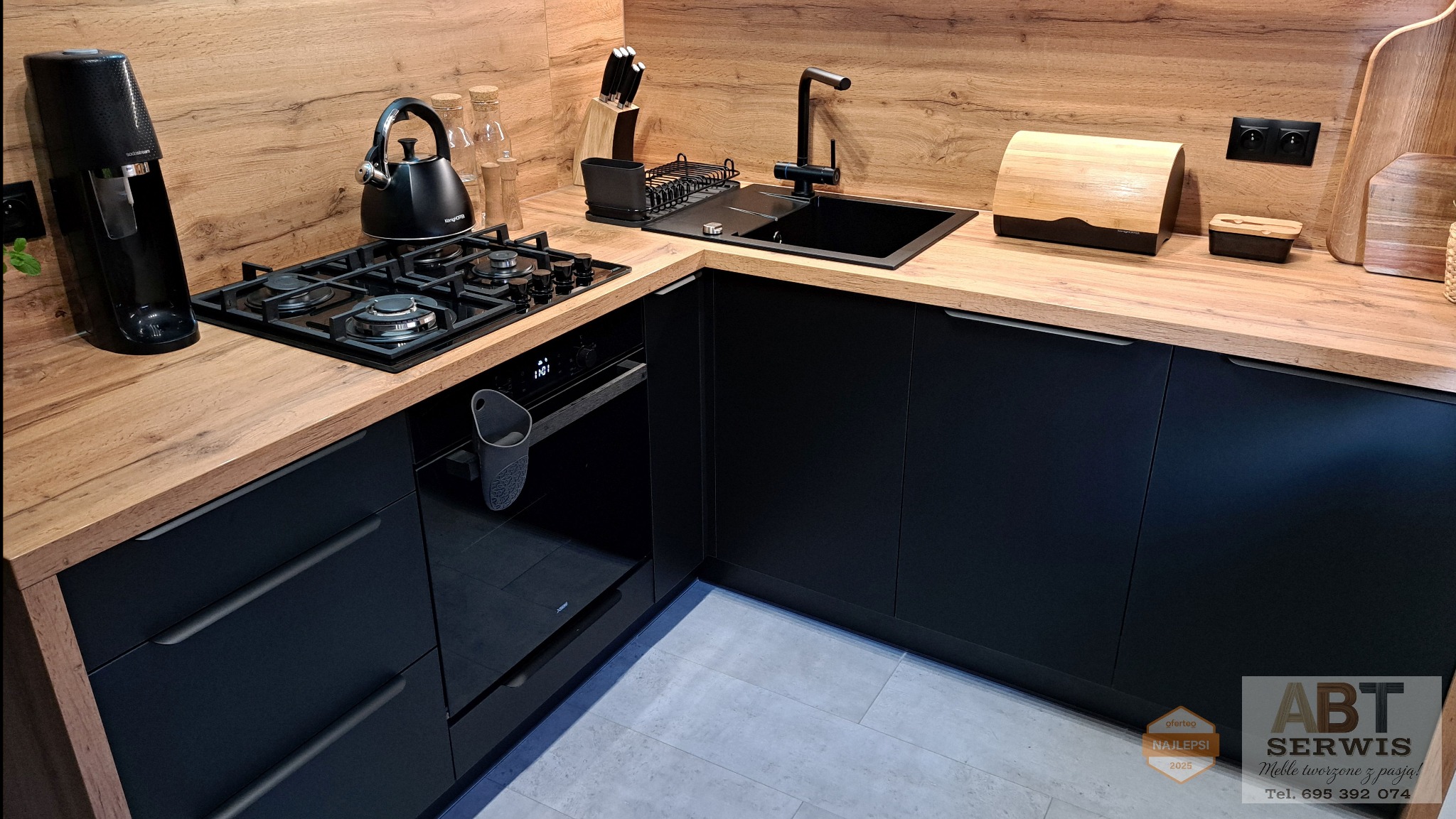 Nowoczesna kuchnia z czarnymi frontami i drewnianym blatem. Widoczny zlew, płyta gazowa, piekarnik i akcesoria kuchenne. Minimalistyczny design, oświetlenie punktowe.