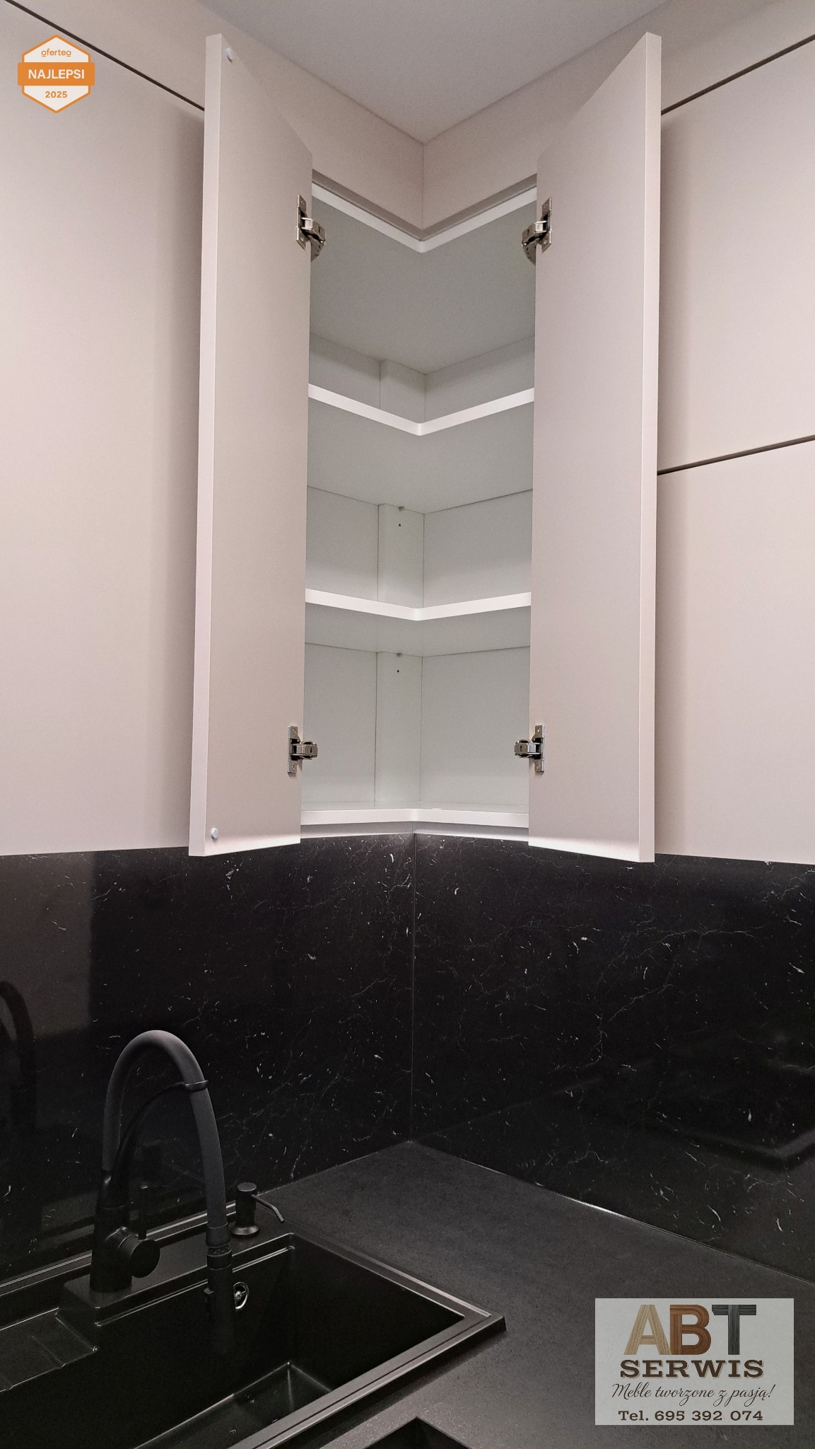 Kremowa szafka narożna z otwartymi drzwiczkami, ukazująca białe półki wewnątrz. Czarny blat z marmurkowym wzorem i czarny zlew. Minimalistyczny design kuchni.